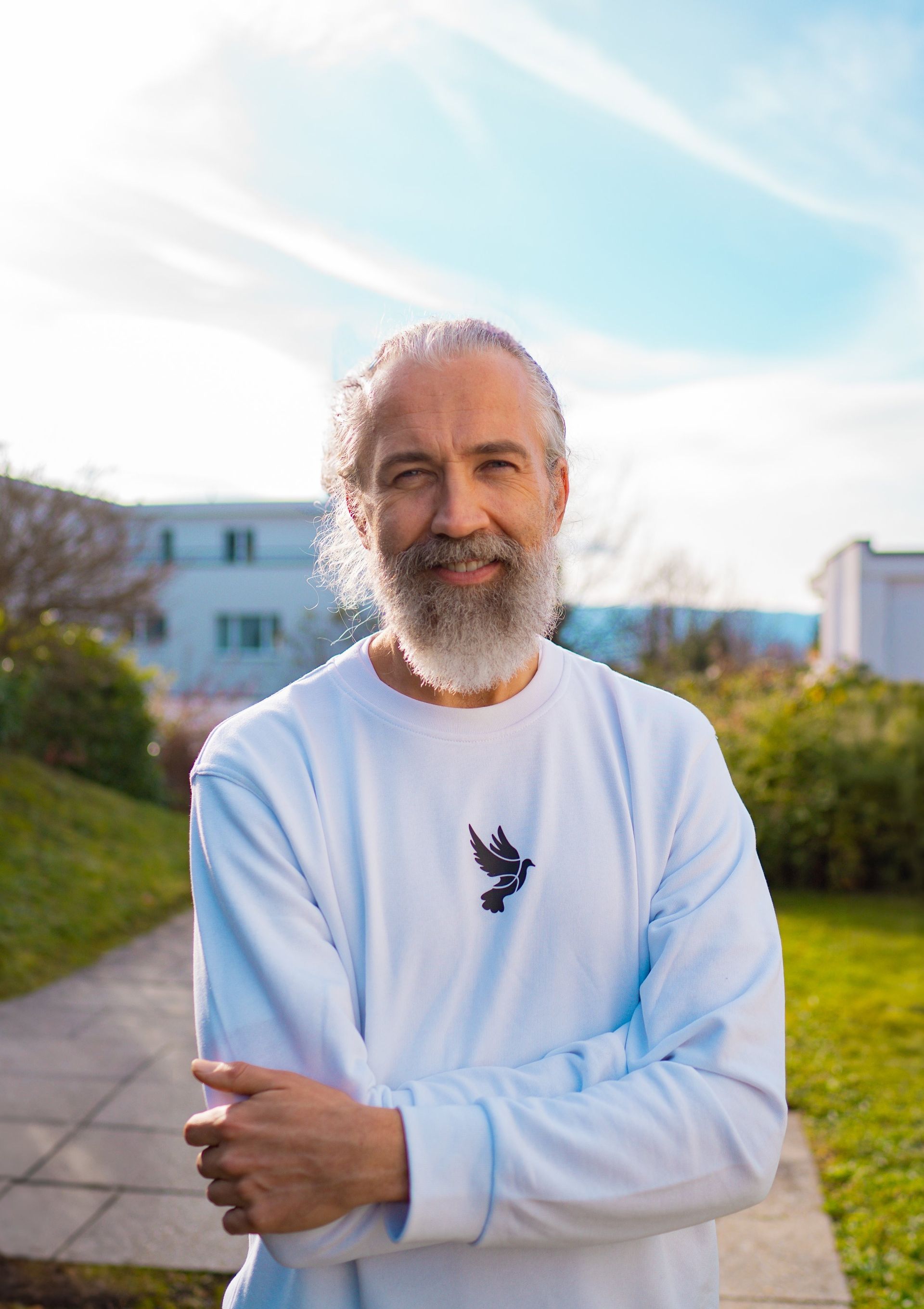 Mann mit langem weißen Haar und Bart im Freien, die Arme verschränkt, trägt ein weißes Hemd.