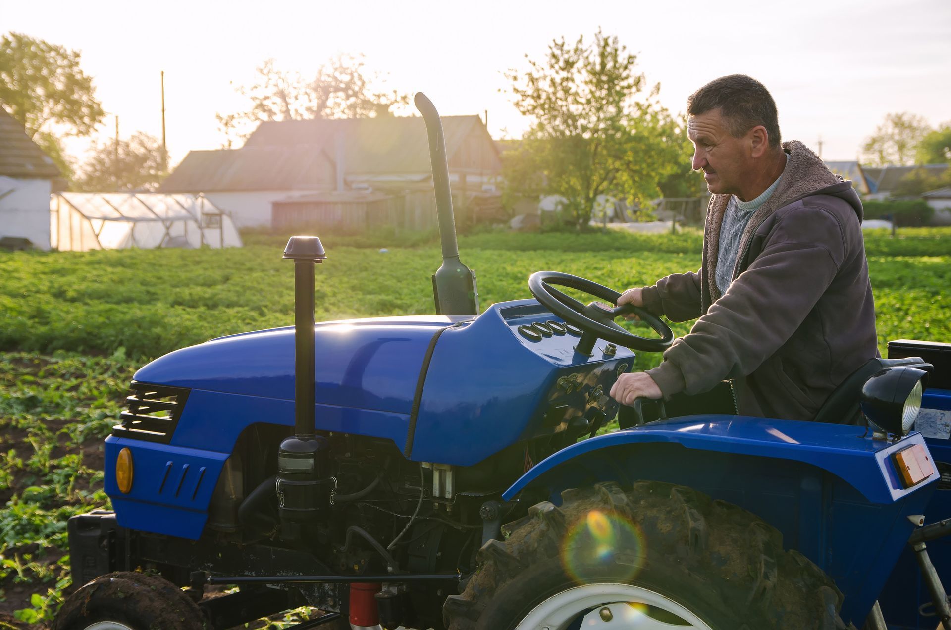 Hombre conduciendo un tractor azul en un campo, iluminado por el sol con casas al fondo.