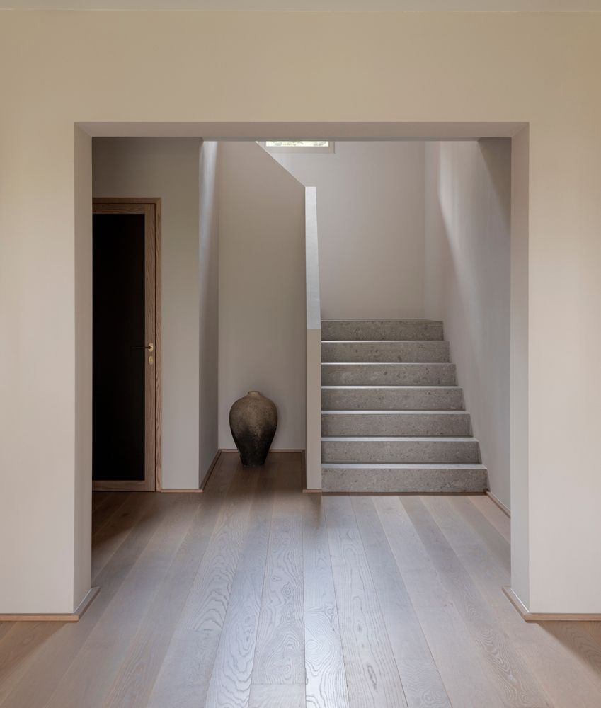 Pasillo minimalista con suelo de madera, puerta y escalera que conduce a un rellano luminoso.