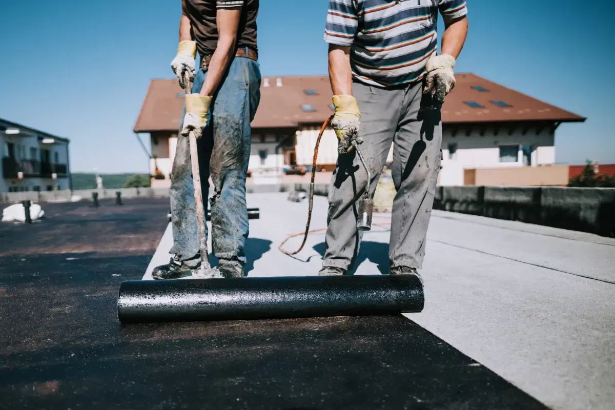 Dos trabajadores extienden una membrana negra para techos sobre un techo plano bajo un cielo soleado.