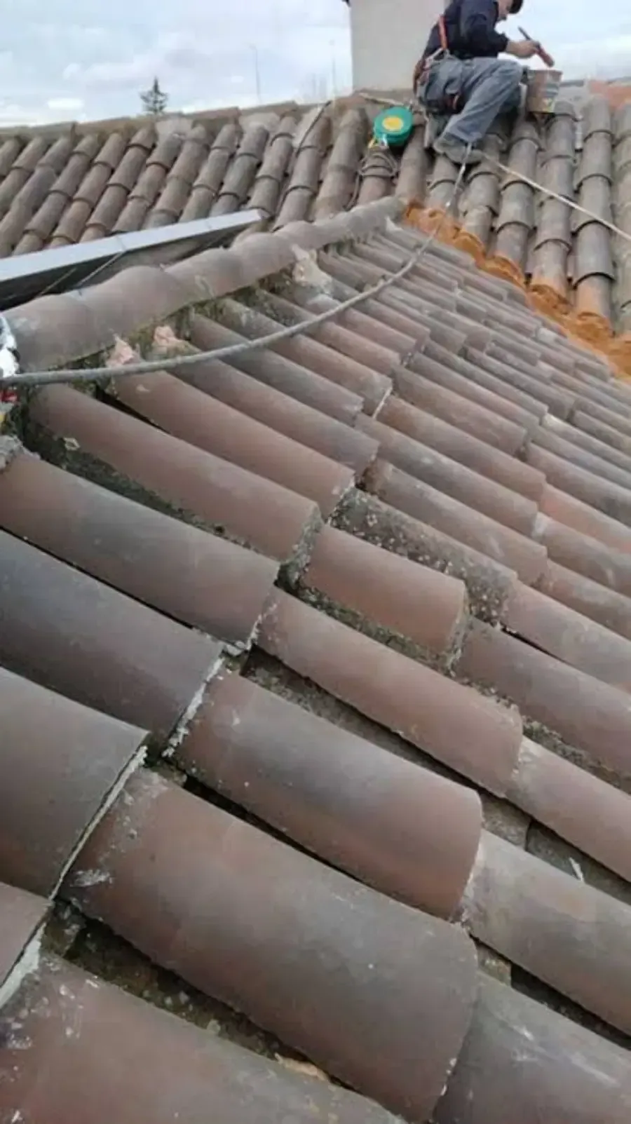 Hombre trabajando en un tejado de tejas de arcilla, probablemente reparando o instalando algo. Cielo nublado.