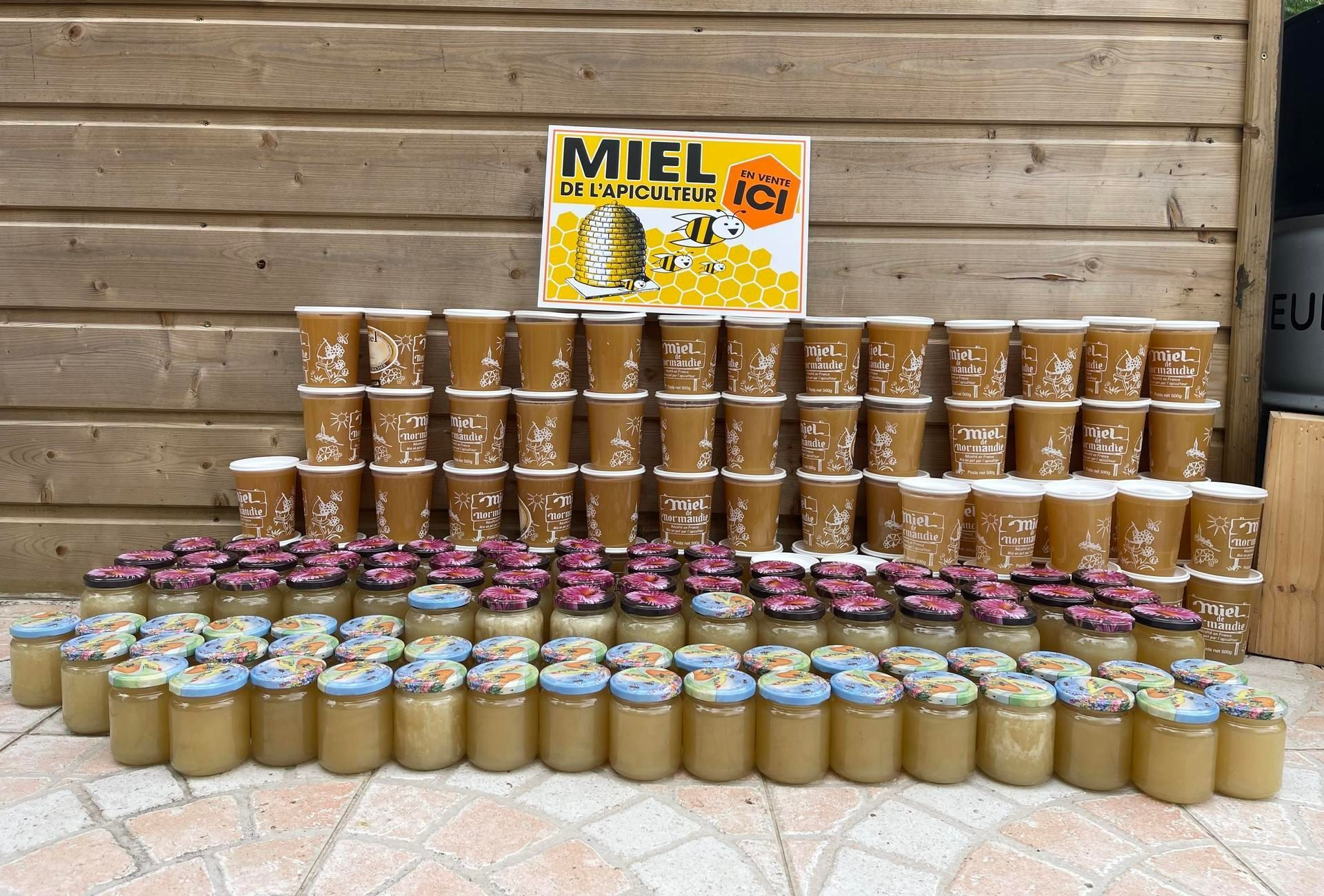 Pots et coupes de miel empilés devant un fond en bois, avec une pancarte.