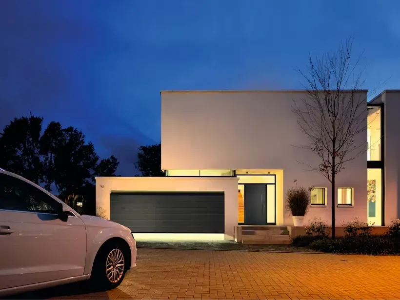 Casa blanca moderna con puerta de garaje gris oscuro, automóvil blanco estacionado y cielo al atardecer.