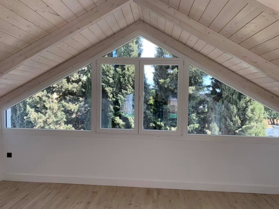 Habitación abuhardillada con ventanas en ángulo, vigas de madera, paredes y suelo blancos, con vistas a árboles verdes.