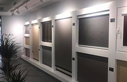 Exhibición de varias persianas para ventanas en una sala de exposición, mostrando diferentes estilos y colores.