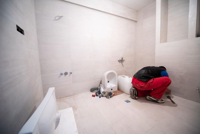 Baño pequeño sin terminar, con paredes alicatadas, un inodoro y un montón de ropa o bolsas en el suelo.