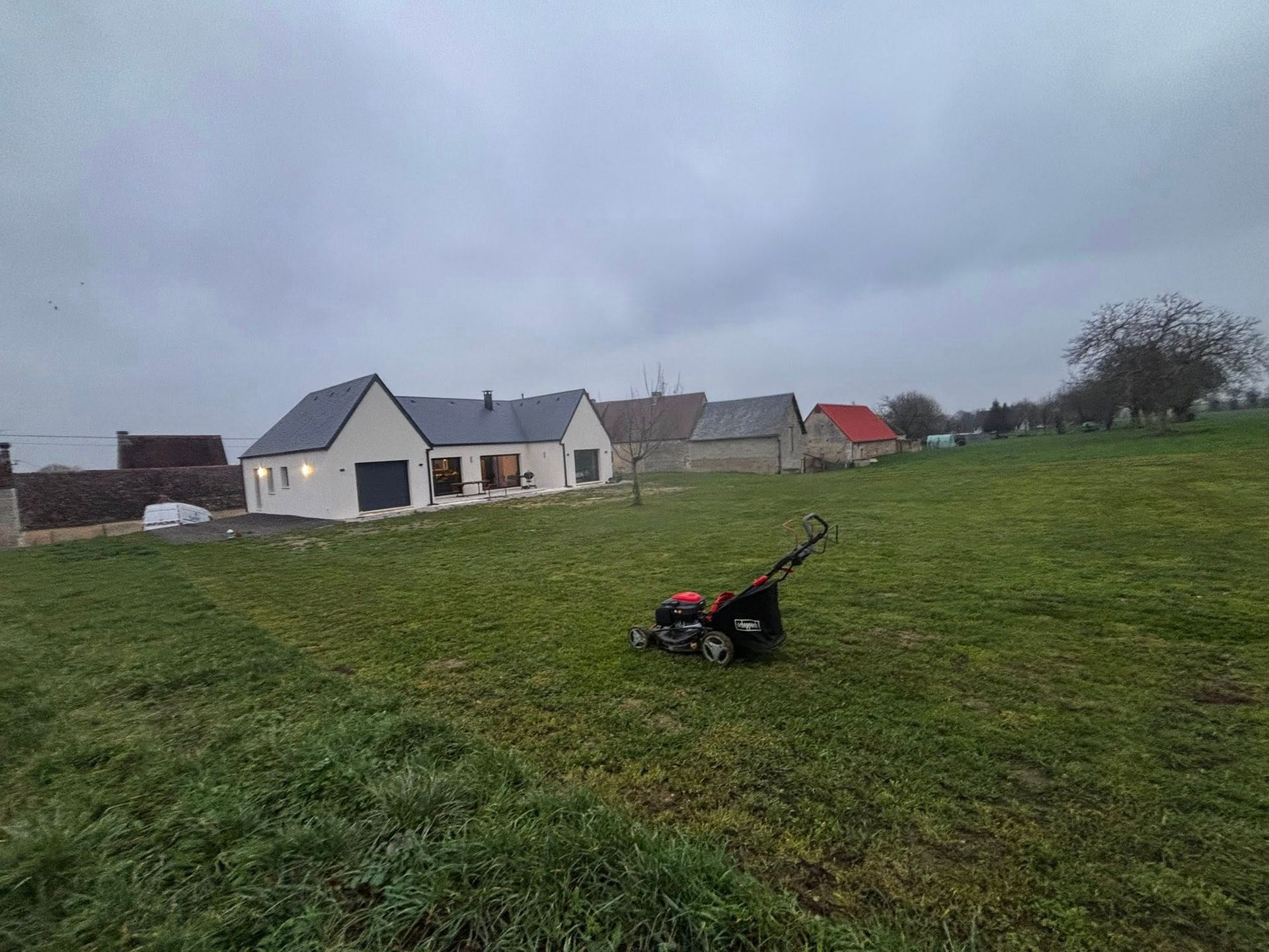 Une tondeuse à gazon est posée dans un champ devant une maison par temps nuageux.