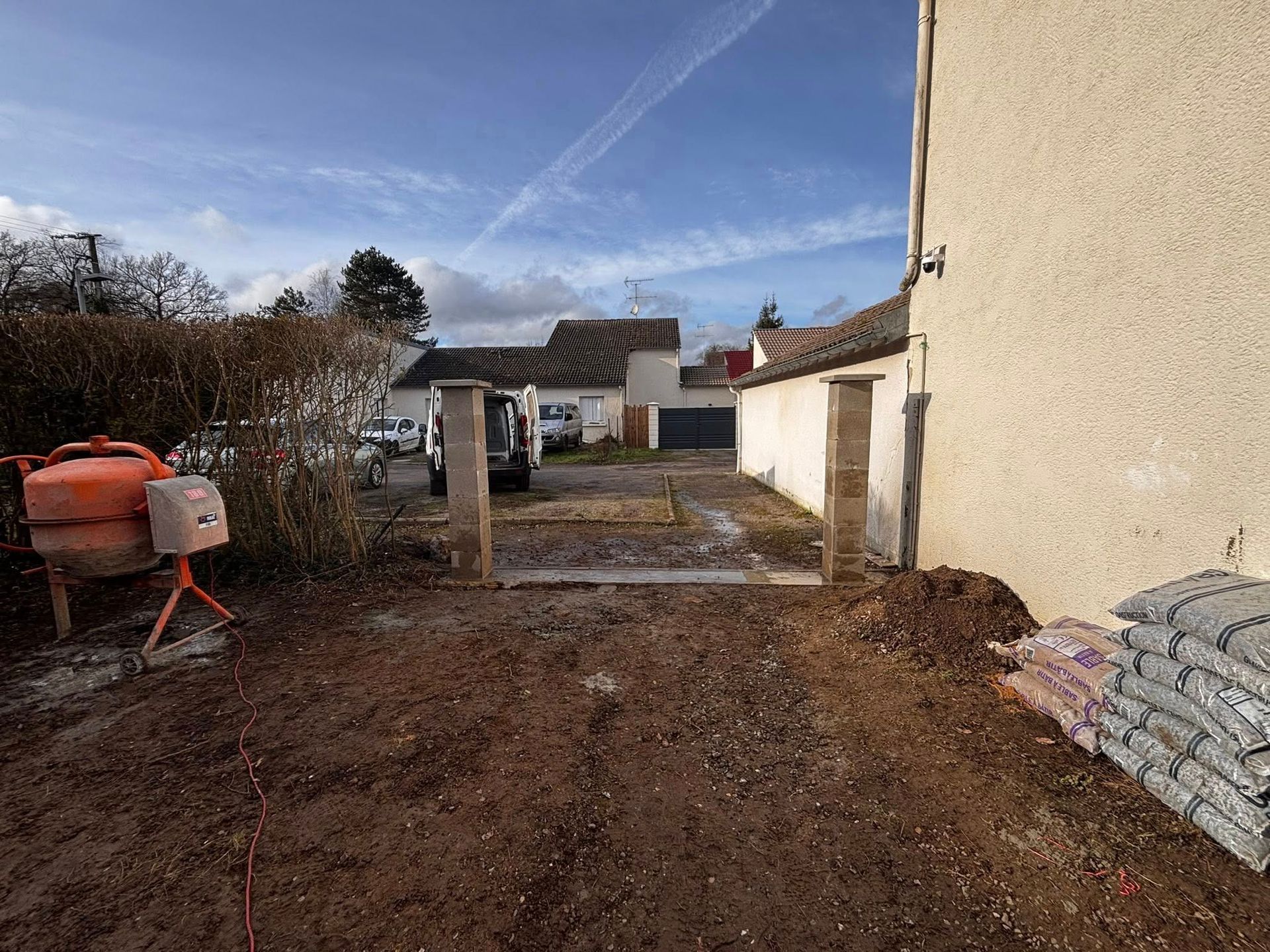 Chantier : zone de terre avec piliers en béton, bétonnière, sacs de matériaux et camionnette stationnée.