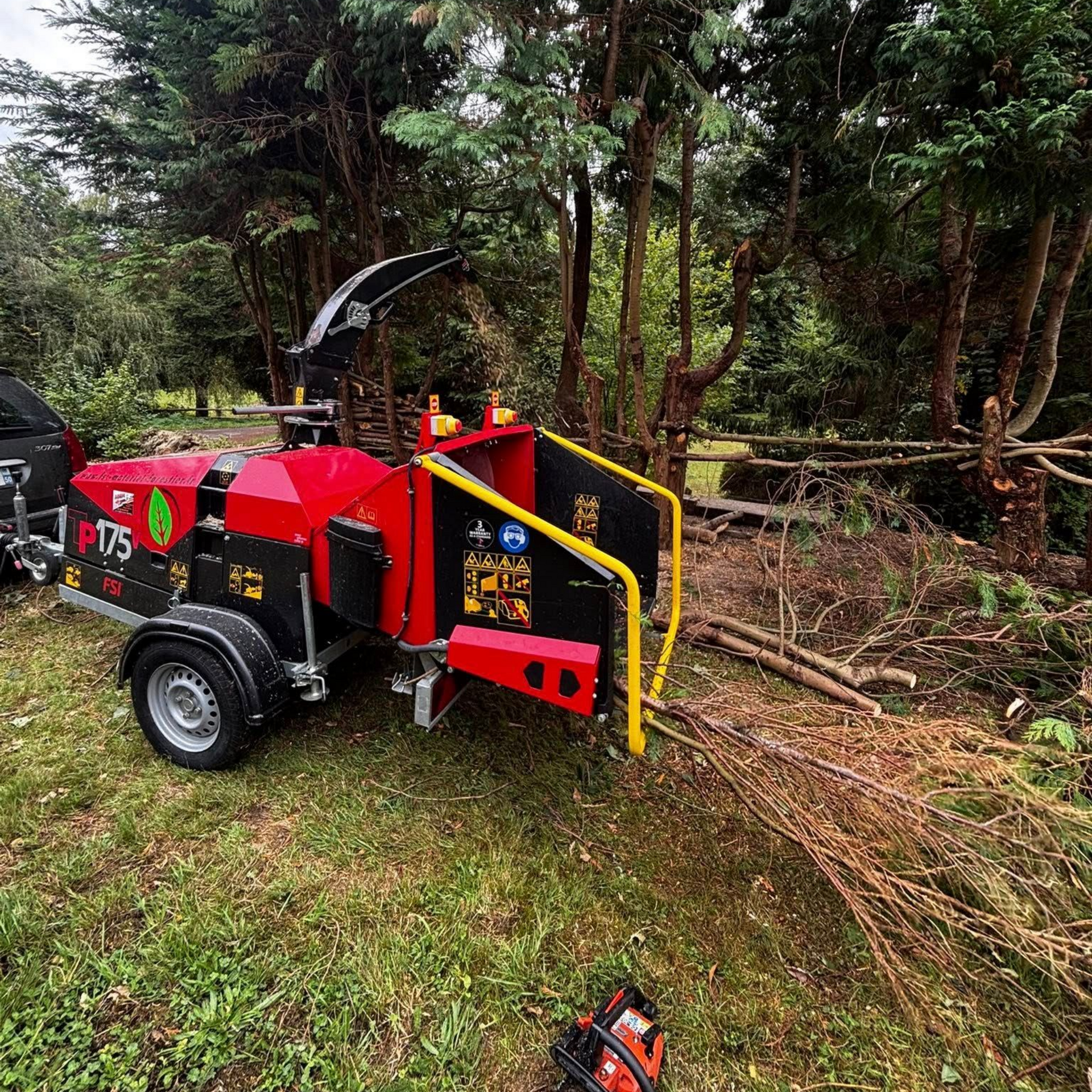 Une remorque à broyeur de branches rouge et noir est garée à côté de grumes dans une forêt. Une tronçonneuse repose au sol.