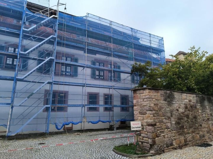 Gebäude im Umbau mit Gerüst und blauem Netz. Steinmauer und Kopfsteinpflasterstraße im Vordergrund.