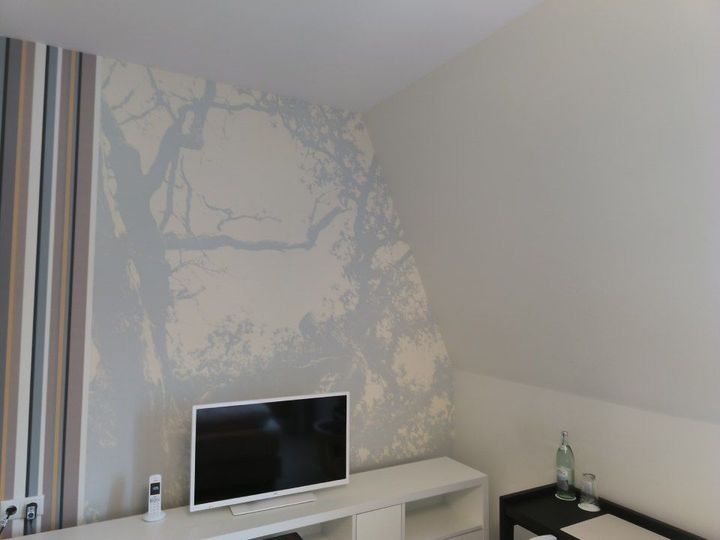 Schlafzimmer mit hellgrauen Wänden, einem Fernseher auf einer weißen Konsole und einem gedeckten blauen Baummotiv an einer Wand.