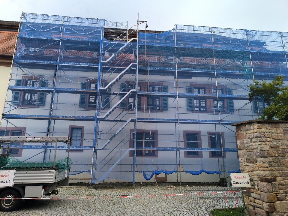 Gebäudefassade im Umbau, mit Gerüsten und blauem Netz verkleidet. Ein weißes Gebäude mit Fensterläden.
