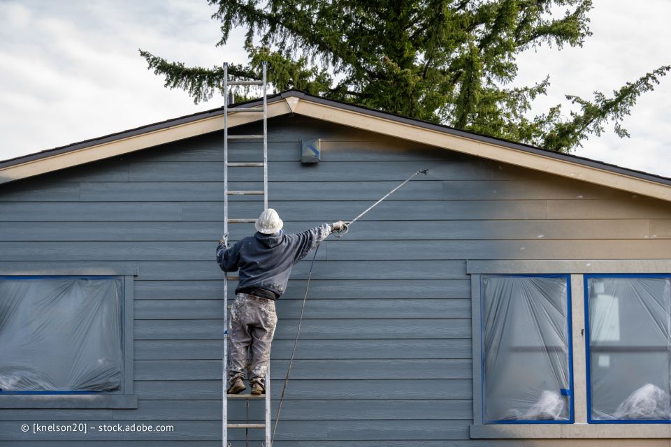 Person auf einer Leiter streicht die blaue Außenverkleidung eines Hauses. Fenster sind abgedeckt. Grüner Baum im Hintergrund.