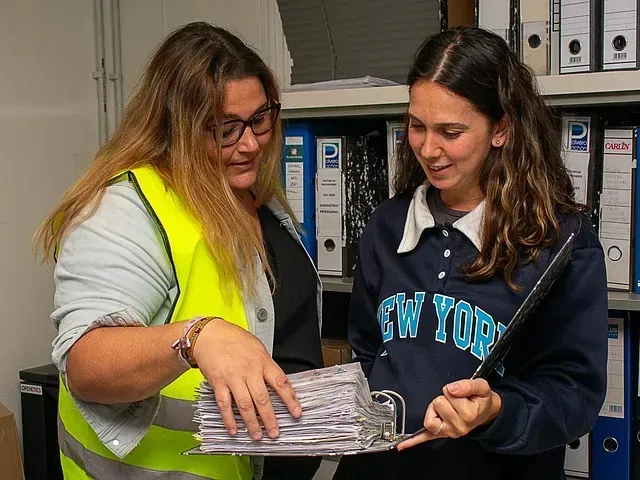 Dos mujeres están paradas una al lado de la otra en una habitación mirando una carpeta.