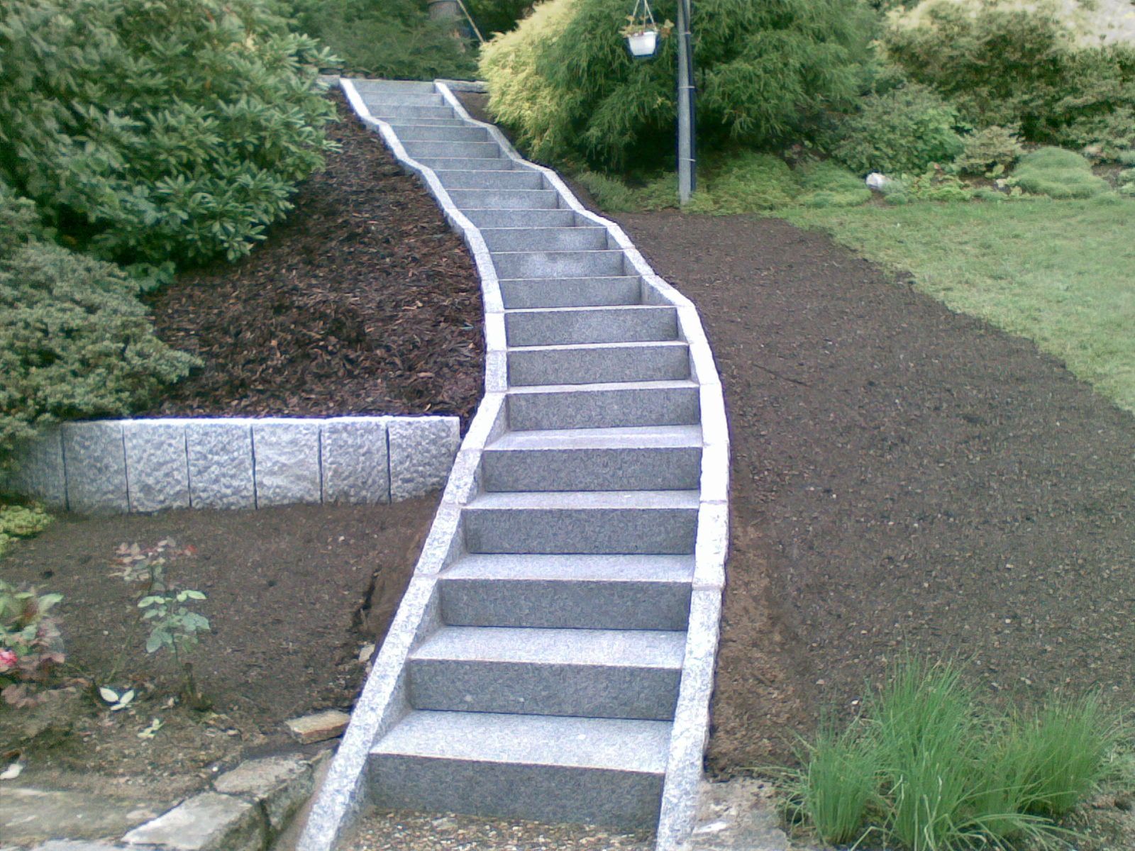 Steintreppe und Stützmauer neben einem Haus, mit grünen Sträuchern und Rasen.