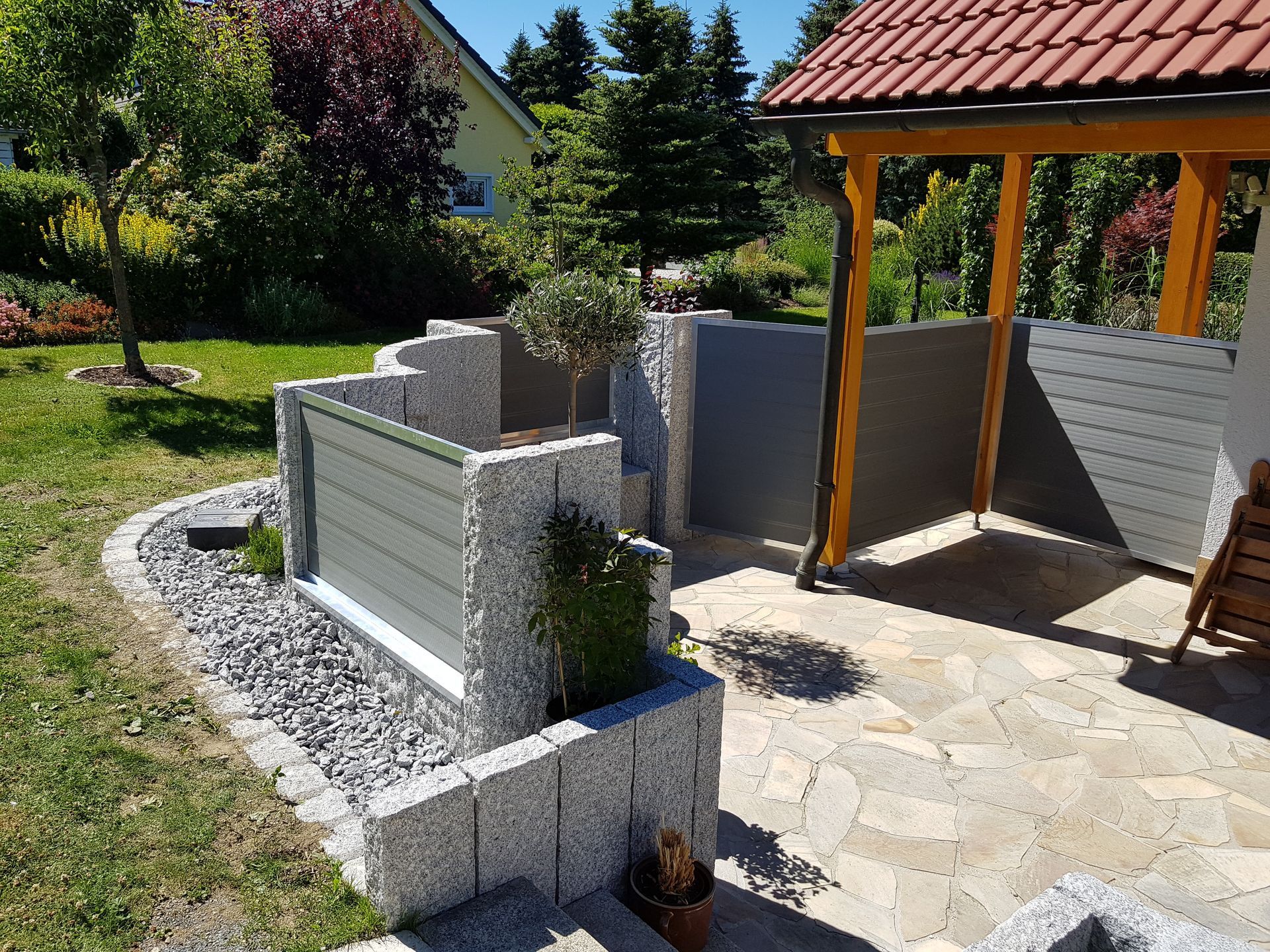 Ein moderner Holzlattenzaun säumt einen sonnigen Gehweg neben hohen Bäumen und Sträuchern.