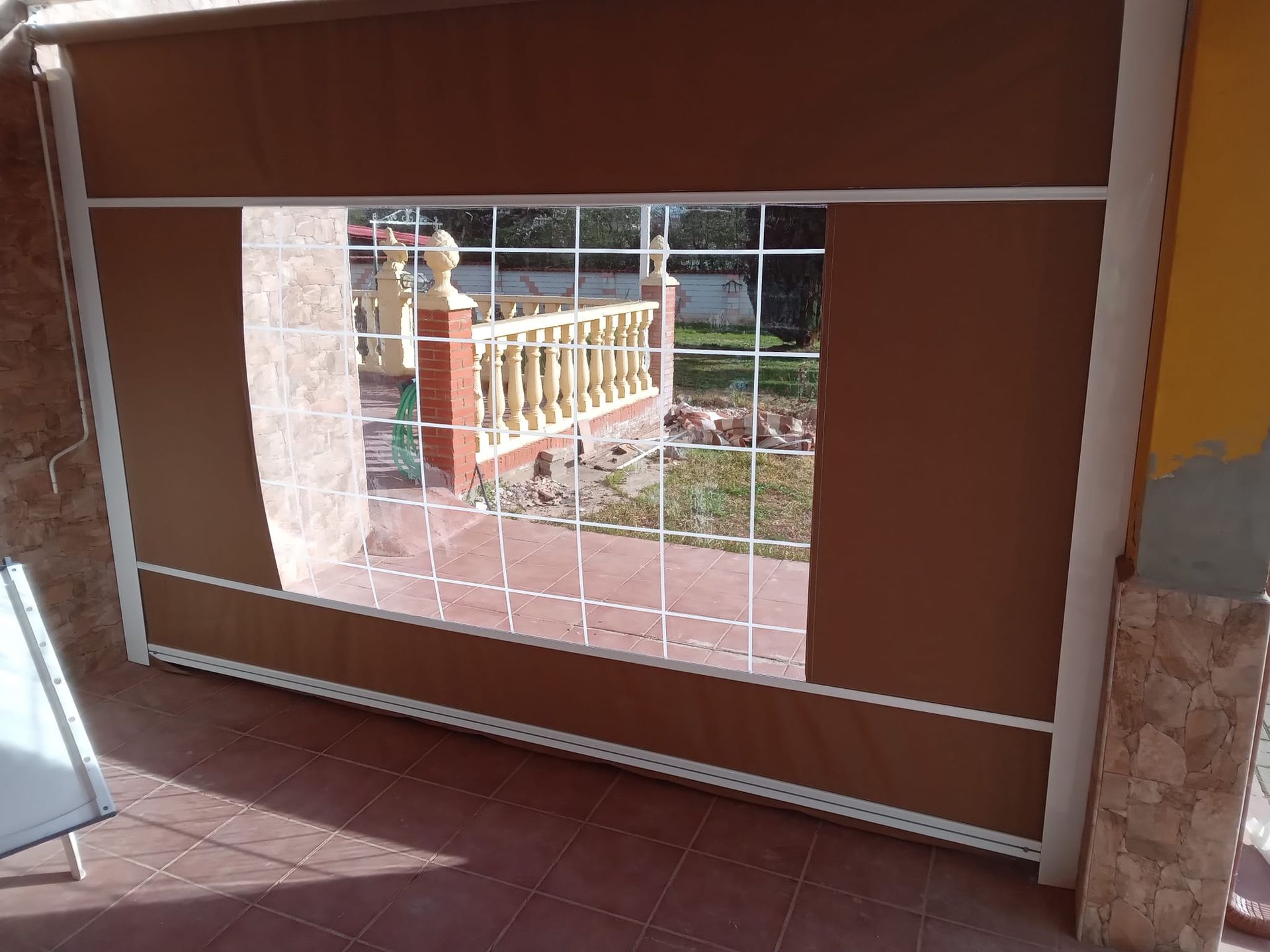 Persianas exteriores marrones que cubren parcialmente una ventana y enmarcan una vista de un porche con barandillas decorativas.