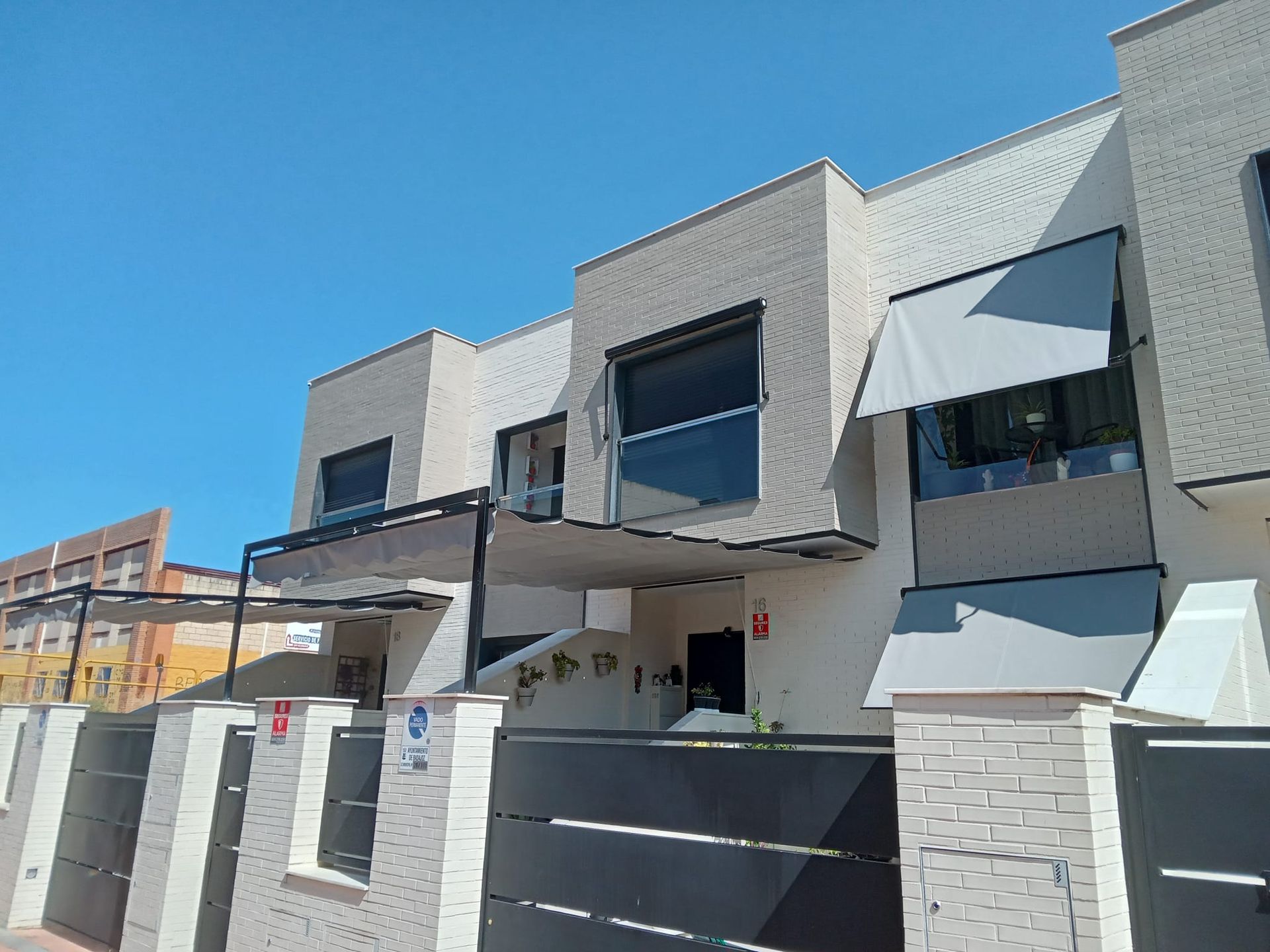 Casas adosadas modernas con exteriores de colores claros, toldos y una puerta negra contra un cielo azul.