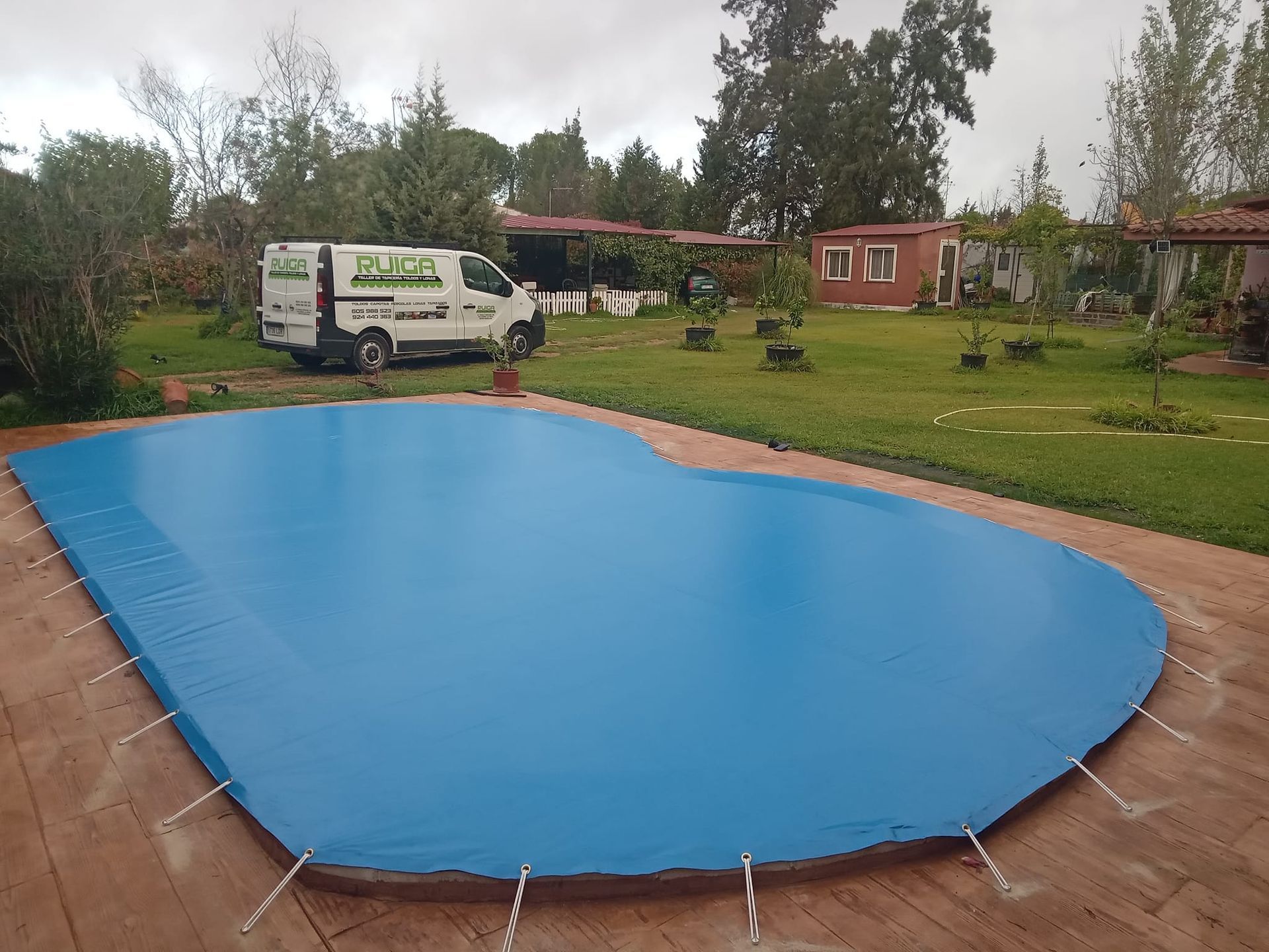 Cubierta de piscina azul sobre una piscina, camioneta al fondo, patio verde, día nublado.