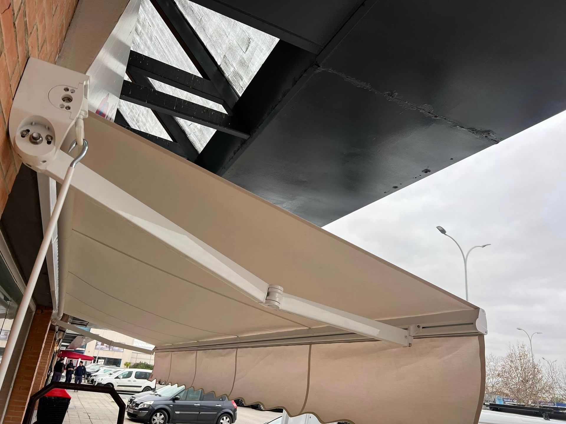 Un toldo beige se extendía desde un edificio. Arriba, una estructura metálica oscura. Coches aparcados cerca.