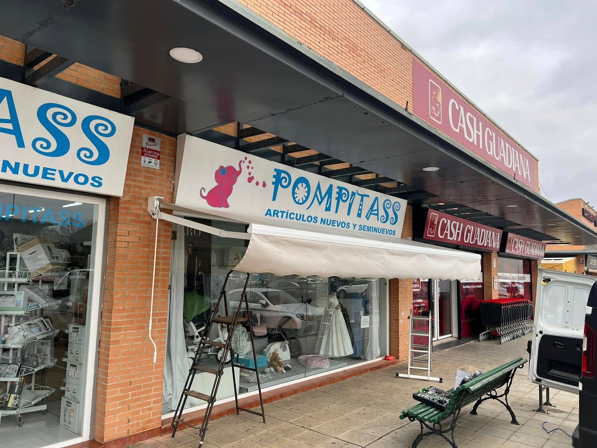 Fachada de tienda con toldo; cartel de Pompitas sobre ventana de tienda, pared de ladrillos rojos, bancos afuera.