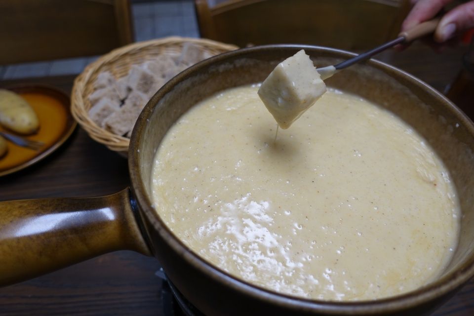 Käsefonduetopf mit einem Brotwürfel auf einer Gabel. Kartoffeln und Brot stehen daneben.
