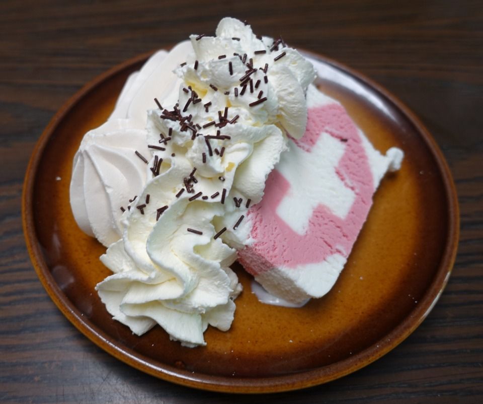 Ein Stück Eistorte mit Schweizer Kreuzmuster, garniert mit Schlagsahne und Schokoladenstreuseln auf einem braunen Teller.