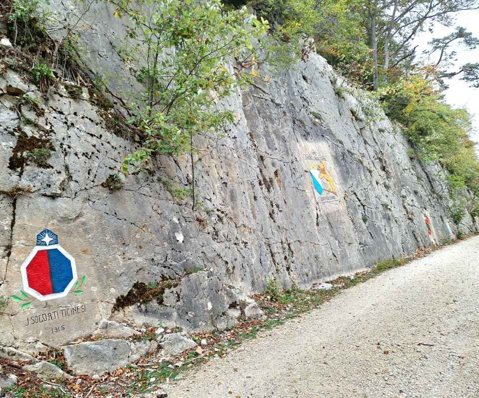 Eine Vase mit bunten Blumen und rosa Schleife steht auf einem Holztisch; im Hintergrund sind eine Bank und eine Mauer zu sehen.
