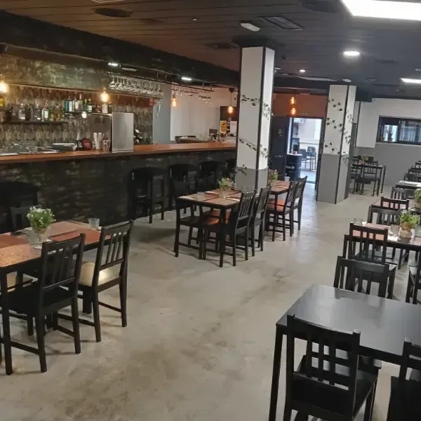Interior de un restaurante con mesas, sillas, barra y taburetes metálicos. Suelos de hormigón, ladrillo visto y una columna con enredaderas.