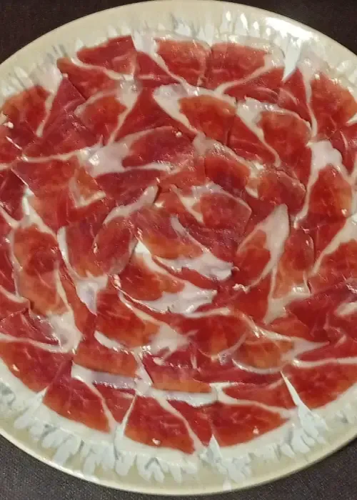 Jamón curado en lonchas finas, presentado en un plato decorativo. Carne roja con veteado blanco visible.