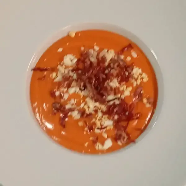 Sopa cremosa de gazpacho con aderezo de huevo y jamón en un tazón blanco.