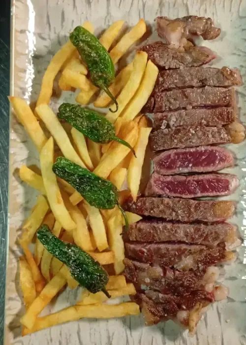 Filete en rodajas con patatas fritas y pimientos verdes fritos en un plato rectangular.