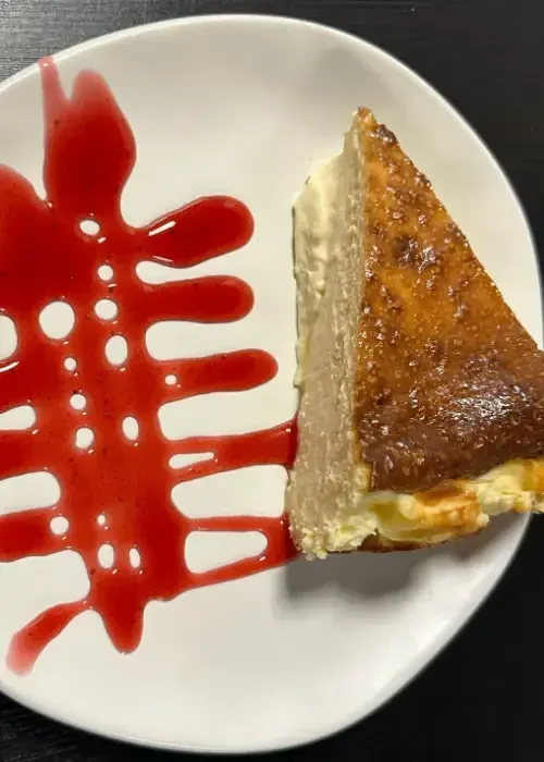 Rebanada de tarta de queso con cubierta caramelizada y un chorrito de salsa roja sobre plato blanco.