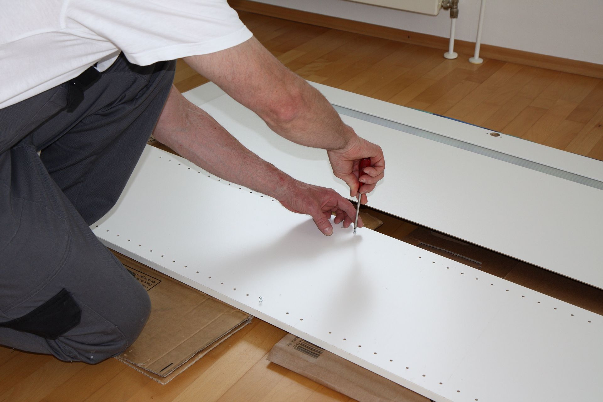 Eine Person montiert kniend mit einem Schraubenzieher weisse Möbel.
