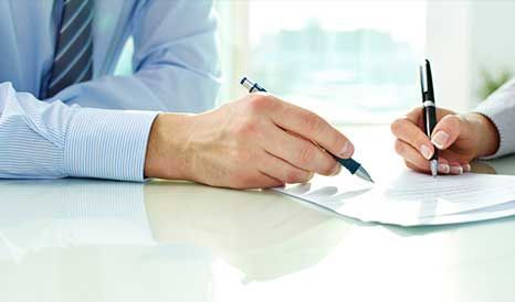 Dos personas firmando un documento en una mesa; primer plano de manos sosteniendo bolígrafos.