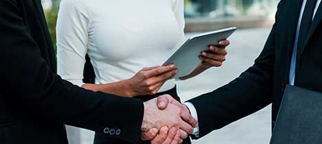 Dos hombres de negocios se dan la mano, al fondo una mujer con camisa blanca sosteniendo una tableta.