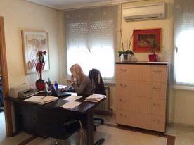 Mujer en un escritorio de oficina, con papeles y portátil. Arte rojo y flores, cajones de madera clara y ventana con persianas.