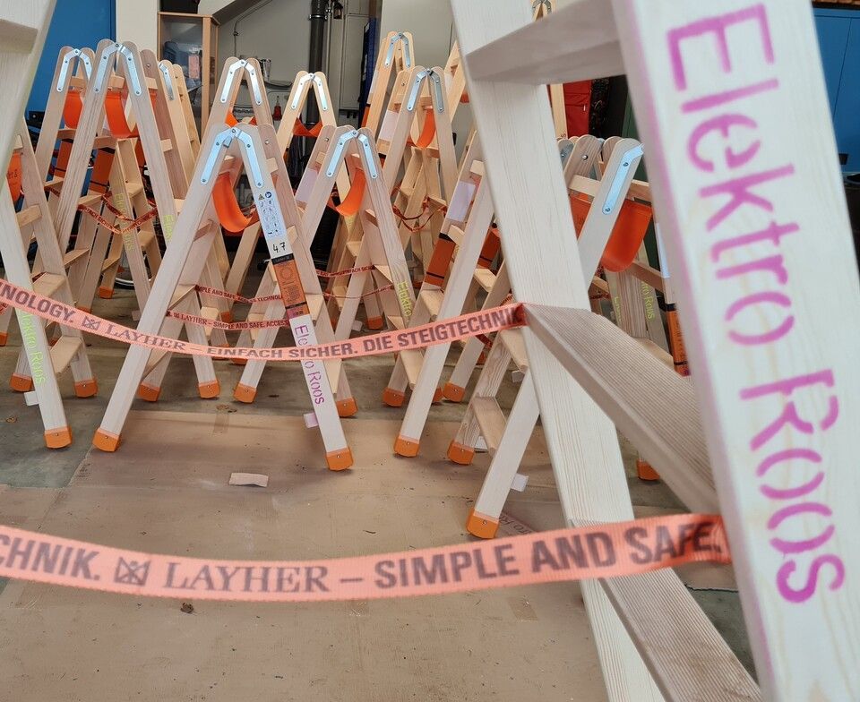 Reihen von Holzleitern in einer Werkstatt; orangefarbenes Klebeband mit Aufschrift davor. „Elektro Roos“ ist auf eine Leiter gedruckt.