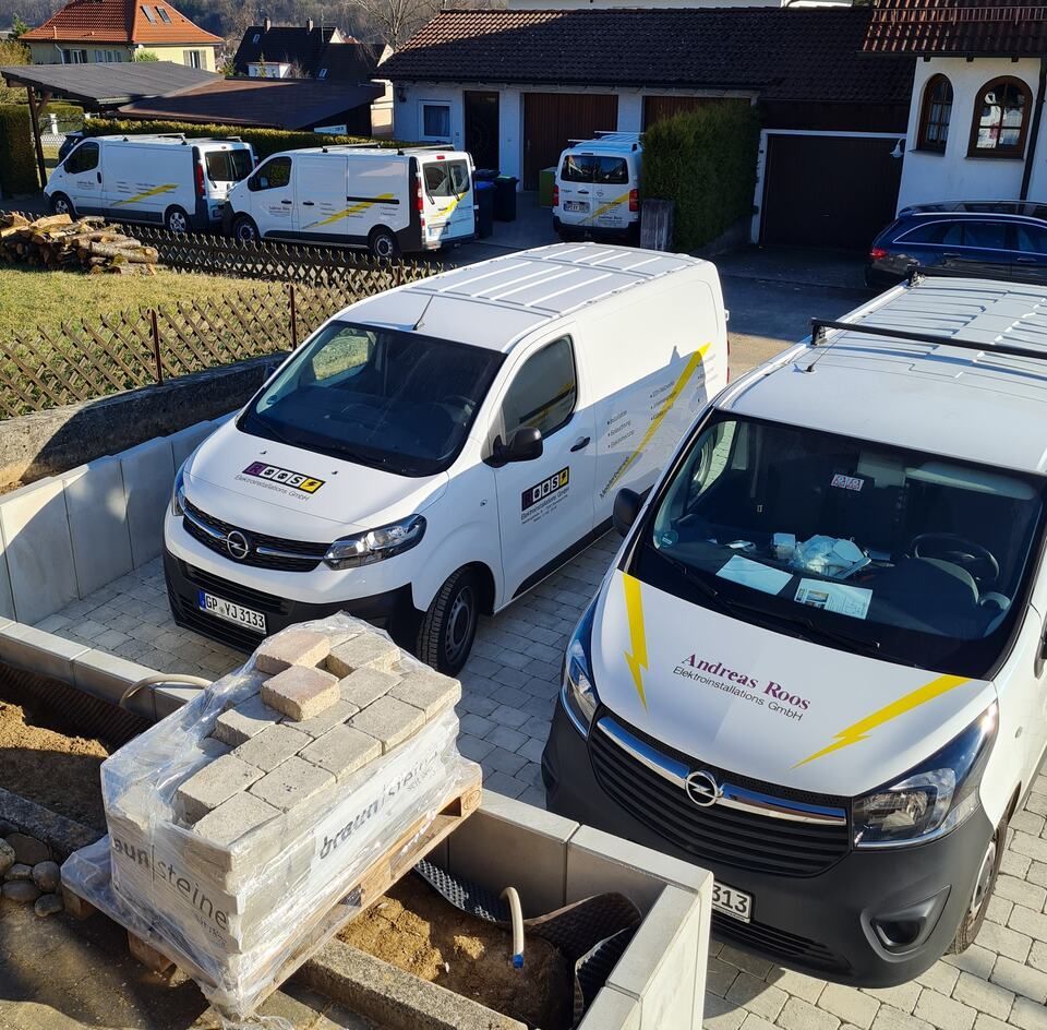 Mehrere weiße Lieferwagen mit Firmenlogo parkten neben einer Baustelle mit Baumaterialien.