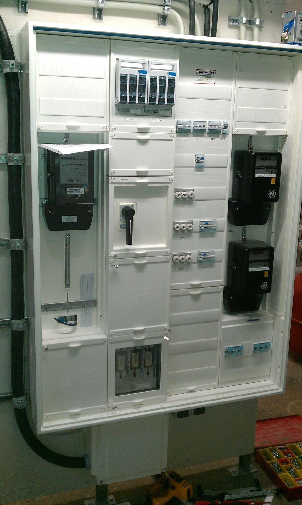 Weißer elektrischer Schaltschrank mit verschiedenen Sicherungsautomaten, Zählern und Schaltern an der Wand.