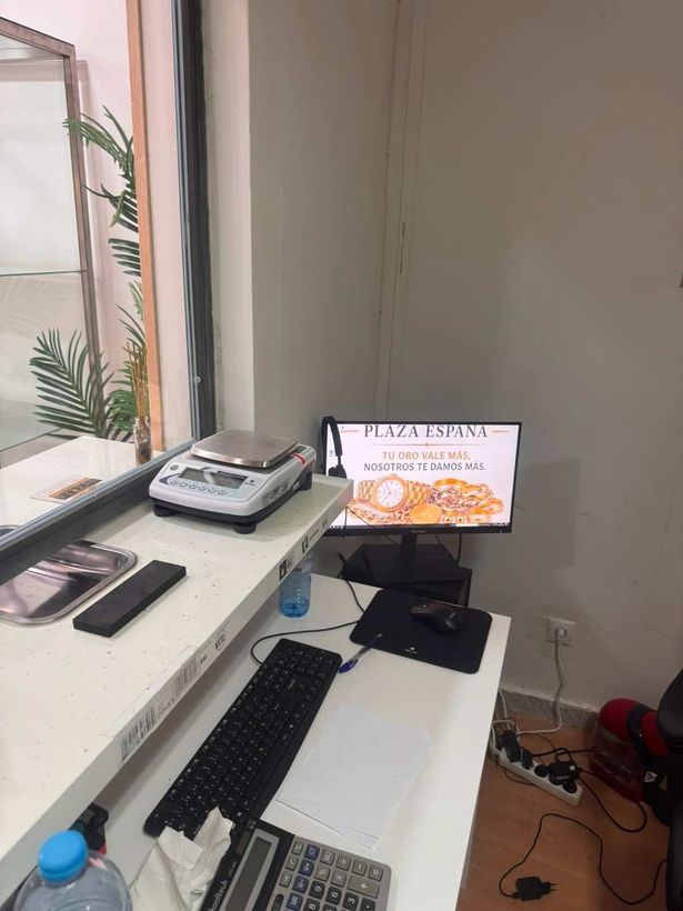 Escritorio con monitor, teclado, teléfono y cartera junto a una ventana, en una pequeña habitación que parece una oficina.