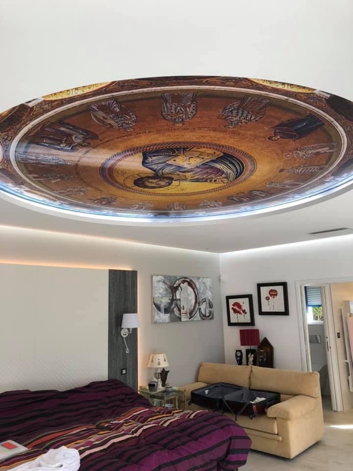 Chambre avec plafond orné d'une fresque, lit, coin salon et œuvres d'art. Tons chauds, lumière naturelle.