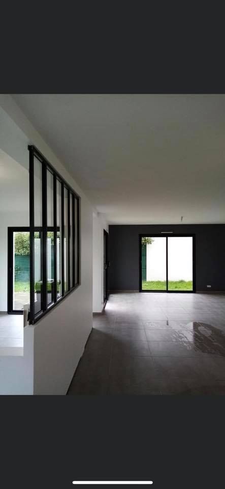 Un intérieur moderne et épuré, avec de grandes fenêtres et une paroi vitrée à cadre noir. Sol et murs gris foncé.