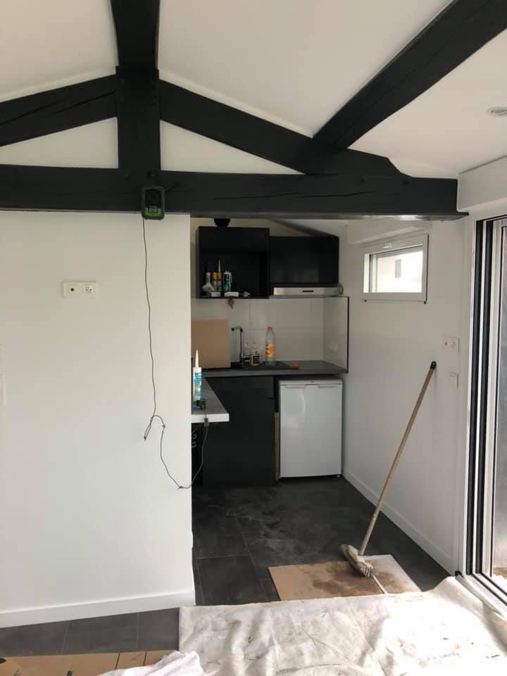 Vue intérieure d'une petite cuisine avec poutres noires, murs blancs et placards gris foncé.