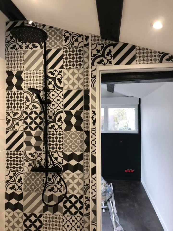 Douche carrelée de carreaux à motifs noirs et blancs avec robinetterie noire ; la porte donne sur une pièce avec un mur sombre.