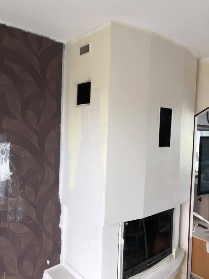 Cheminée blanche avec conduit et fenêtres noirs ; papier peint marron à gauche.