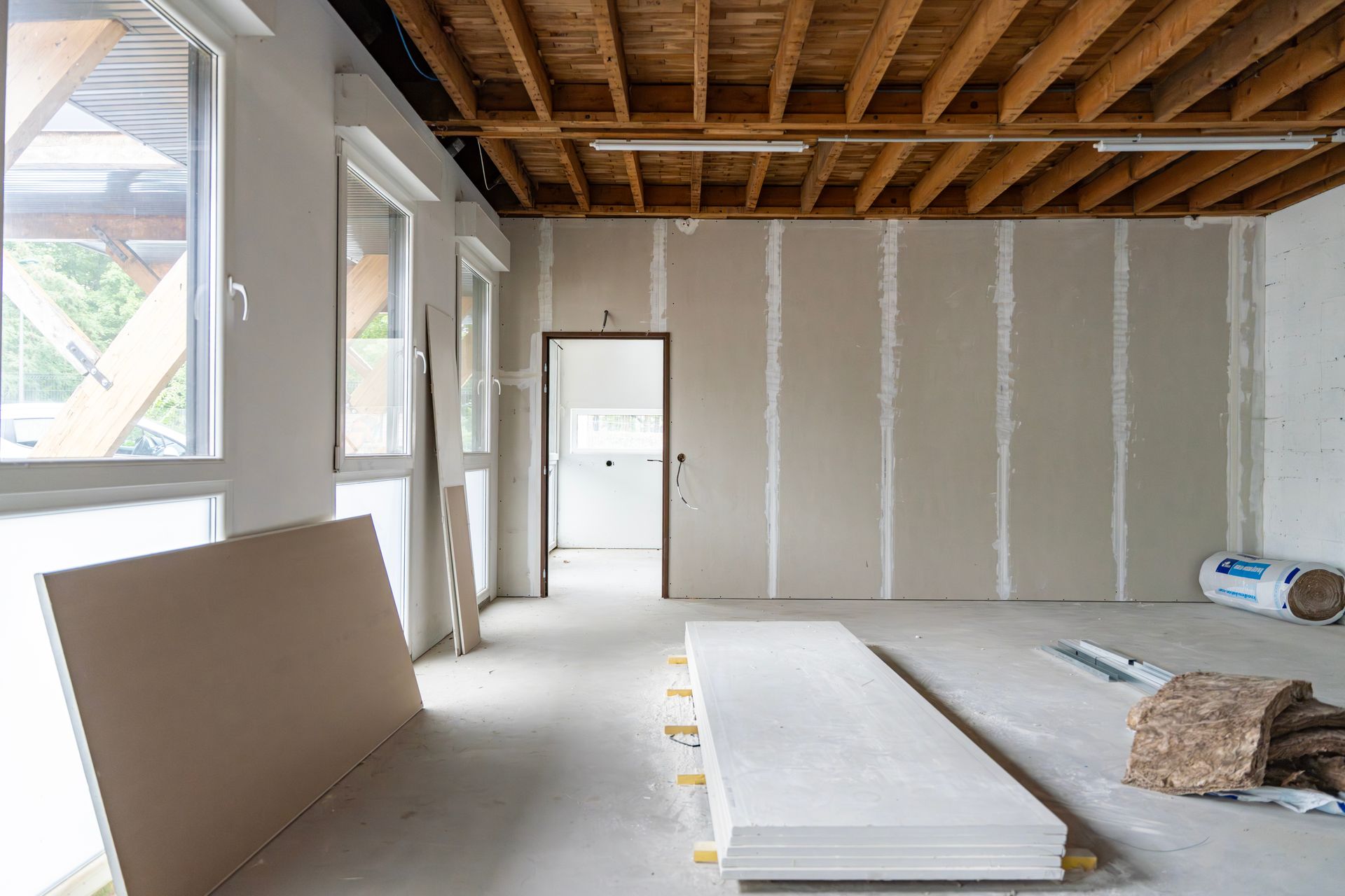 Aménagement intérieur : pièce avec poutres apparentes au plafond, cloisons sèches aux murs, fenêtres et différents matériaux au sol.