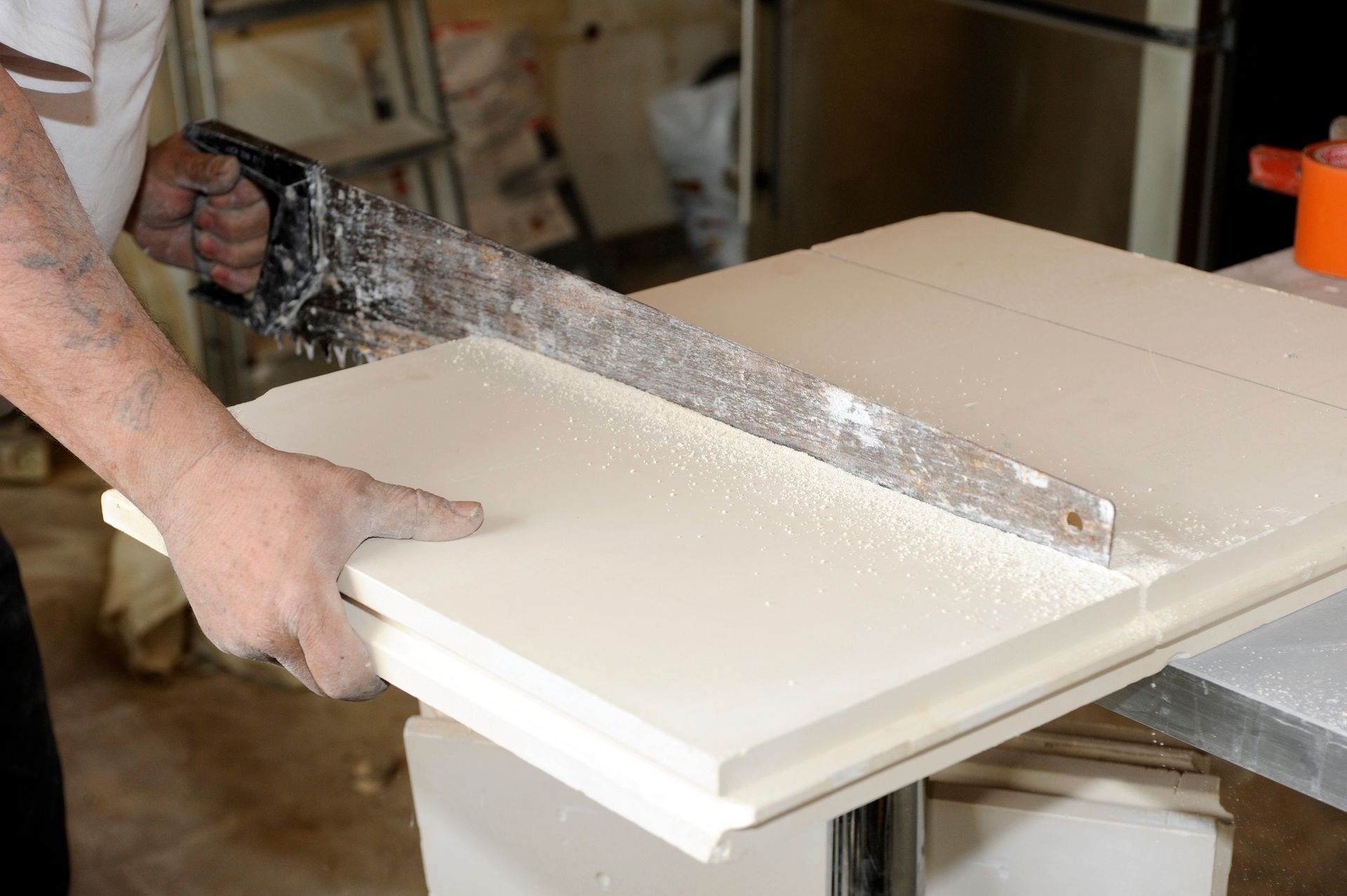 Une personne utilise une scie pour couper un matériau de construction blanc et rectangulaire, créant ainsi de la poussière blanche.