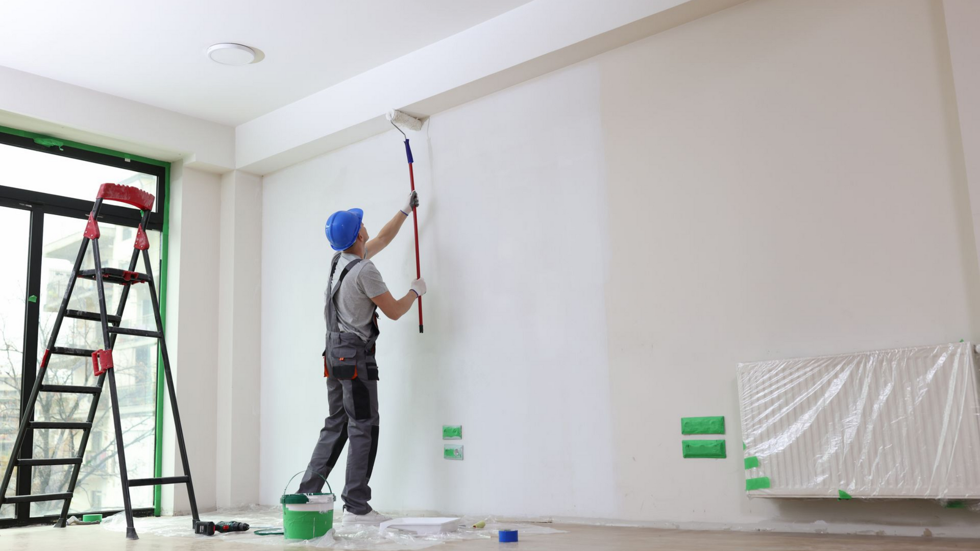 Un peintre portant un casque de chantier bleu utilise un rouleau pour peindre un mur blanc à l'intérieur, une échelle se trouvant à proximité.