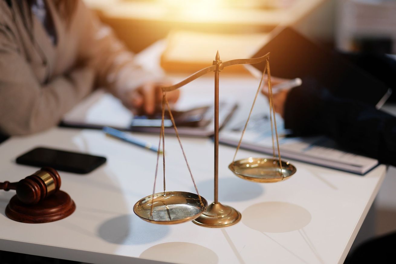 Une balance de la justice sur une table avec un maillet, et des gens qui discutent de documents administratifs.